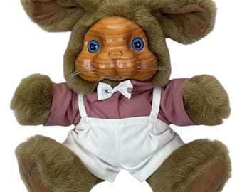 May include: A plush rabbit doll with a brown faux fur hood and body, featuring a smooth, light brown wood-grain textured face with blue eyes and whiskers. The doll wears a mauve collared shirt, a white bowtie with small patterns, and white overalls.