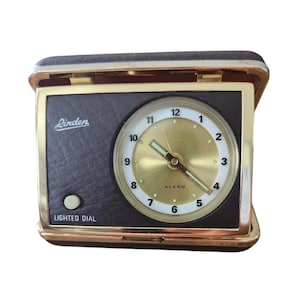 May include: A vintage Linden travel alarm clock. The clock has a gold-tone face with black numbers and hands. The clock is encased in a black and gold folding case. The words "Linden" and "LIGHTED DIAL" are visible on the clock.