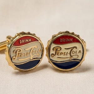 May include: A pair of gold-toned cufflinks featuring the Pepsi-Cola logo. Each cufflink is round, resembling a bottle cap, with red, white, and blue enamel. The word "DRINK" is at the top, and "Pepsi-Cola" is in the center.