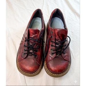 May include: A pair of burgundy leather shoes with black laces. The shoes have a classic design with visible stitching and a rounded toe. The soles are a contrasting brown color. The shoes are displayed on a white surface.
