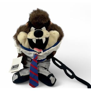 May include: A plush Taz the Tasmanian Devil toy wearing a white shirt, gray pants, and a red and blue striped tie. The toy has a black mask and a tag from Warner Bros. Studio Store.