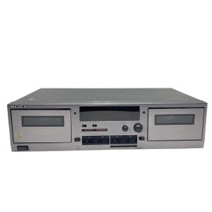 Puede incluir: Una pletina de casete Sony plateada con dos compartimentos para cintas, una pantalla central y botones de control. La pletina tiene un diseño elegante y rectangular, con el logotipo de Sony en la parte superior izquierda.
