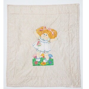 Op de afbeelding: Een vierkante doorgestikte deken met een herhalend beige en wit bloemenpatroon. In het midden staat een grafische illustratie van een pop met oranje haar en roze strikken, gekleed in een witte jurk, staand in een veld met kleurrijke bloemen.