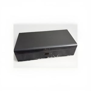 Puede incluir: Una caja rectangular negra con tapa con bisagras. La caja tiene un acabado mate y una pequeña cerradura de combinación en la parte delantera. La caja está sobre una superficie blanca.