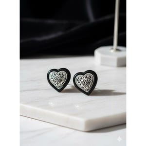 Peut inclure: Une paire de boucles d'oreilles en forme de cœur avec un contour noir lisse et une incrustation centrale argentée ornée de motifs à volutes complexes en relief. Les boucles d'oreilles présentent une finition polie et un design symétrique.