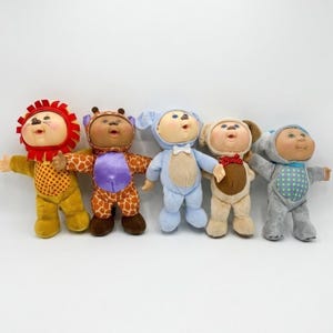 May include: Five Cabbage Patch Kids dolls dressed in animal costumes. The costumes include a lion, giraffe, lamb, dog, and a bear. The dolls are standing in a row with their arms outstretched.