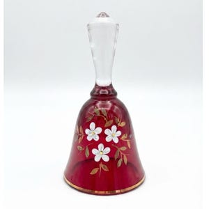 Puede incluir: Una campana de cristal rojo rubí con un mango de cristal transparente y un motivo floral pintado a mano. El diseño incluye tres flores blancas con centros amarillos rodeadas de delicadas hojas y enredaderas doradas, con un borde dorado.