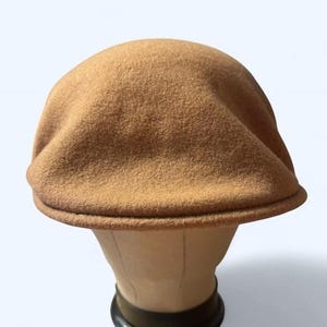 Puede incluir: Gorra plana de color marrón claro, hecha de una tela texturizada. La gorra tiene una corona redondeada y una pequeña visera curvada. El sombrero se muestra en una cabeza de maniquí, mostrando su forma y diseño. Este estilo clásico es adecuado para varias ocasiones.
