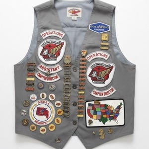 Op de afbeelding: Een grijs vest versierd met talloze geborduurde patches en metalen pins. De patches bevatten teksten zoals Operations, Assistant Chapter Director en Gold Wing Road Riders Association. Een kleurrijke kaart van de Verenigde Staten is ook zichtbaar.