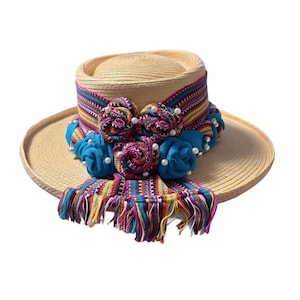 May include: A straw hat with a wide brim and a cylindrical crown. The hat is adorned with a colorful woven band, blue fabric flowers, and a fringed scarf. Small white pearls are also attached to the hat.