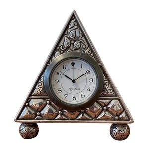 Puede incluir: Un reloj de escritorio triangular plateado con una esfera redonda. La esfera es blanca con números y manecillas negras. El reloj está sostenido por dos pies esféricos decorativos. La palabra "Brighton" está impresa en la esfera.