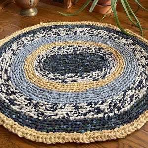 May include: Oval braided rug with concentric circles of color. The rug features a light beige border, followed by a light blue, beige, and dark blue center. The rug is made of fabric strips and is suitable for a living room or bedroom.