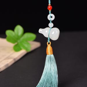 Peut inclure: Pendentif blanc en forme de gourde avec un pompon turquoise et une perle rouge. Le pendentif est enfilé sur un cordon turquoise avec un accent doré. Le numéro "13287" est visible dans le coin supérieur gauche.