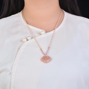Puede incluir: Un collar con colgante rosa claro con diseño de nube. El colgante cuelga de un cordón beige con detalles rosas y cuentas doradas. El collar se lleva sobre una blusa blanca con botones perlados.