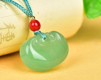 May include: A light green jade pendant in a rounded shape, suspended from a teal cord. A small red bead and gold-colored accent sits above the pendant. The pendant has a smooth, polished surface and is likely a piece of jewelry.