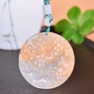 May include: A round pendant with a swirling, cloud-like design in shades of cream, orange, and gray. The pendant is suspended from a teal cord with white and teal beads. The pendant has a polished, smooth surface.