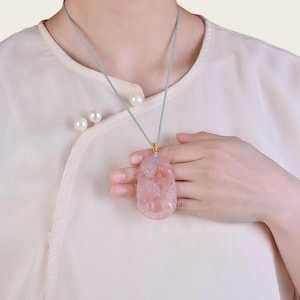 Op de afbeelding: Een roze hangerketting met een gesneden ontwerp. De hanger hangt aan een lichtblauw koord met een gouden sluiting. De ketting wordt gedragen over een crèmekleurige blouse.