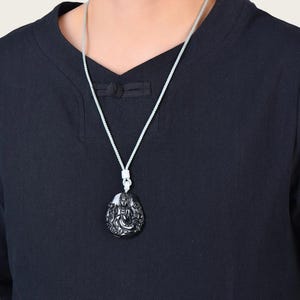 Può includere: Una collana con pendente nero con un'intagliatura dettagliata di una figura seduta. Il pendente è appeso a un cordino bianco. La persona indossa una camicia nera.