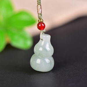 Puede incluir: Un colgante de jade con forma de calabaza, de color verde pálido, suspendido de un cordón verde con una cuenta roja y detalles dorados. La forma de la calabaza es un símbolo tradicional de salud y longevidad.