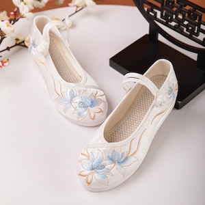 以下が含まれることがあります： 青い花柄の刺繍が施された白いフラットシューズ。靴の上部にストラップがあり、小さなバックルが付いています。