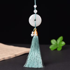 Könnte beinhalten: Ein weißer Jade-Anhänger mit einem zentralen Loch, verziert mit einer hellblauen Quaste und kleinen Perlen. Der Anhänger wird von einer passenden Kordel gehalten und ist ein elegantes Accessoire. Das Design beinhaltet goldene Akzente.
