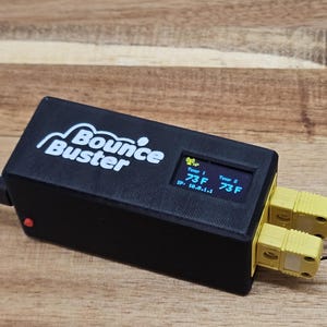May include: Black electronic device with the text "Bounce Buster" in white. The device has a small screen displaying temperature readings in Fahrenheit, and two yellow connectors. A red button is visible on the side.