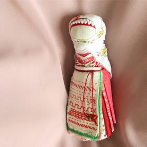 May include: A handmade fabric doll wearing a white head covering and a red and white dress with green and white embroidery.