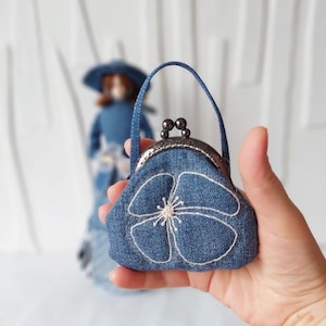 Pode incluir: Uma pequena bolsa de ganga azul com um desenho de flor bordada branca e um fecho prateado. A bolsa tem uma alça preta.
