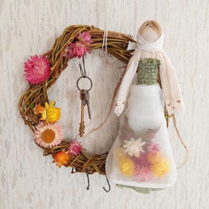 Puede incluir: Una muñeca de tela blanca con un corpiño verde y un pañuelo blanco en la cabeza cuelga de una corona de ramas decorada con flores secas. La muñeca sostiene una bolsa de tela transparente llena de flores secas.