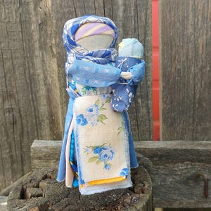 以下が含まれることがあります： 青い花柄の布を着た手作りのかやの人形。人形は青い布で包まれた小さな人形を抱いています。