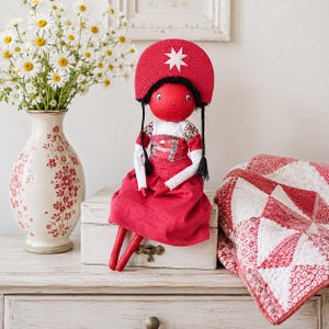 May include: A handmade fabric doll with a bright red face and matching red headpiece featuring a white star. The doll wears a white long-sleeved top with floral accents and a solid red skirt. It has black braided hair and sits upright.