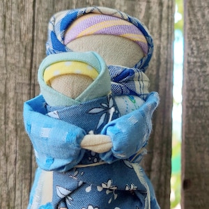 Op de afbeelding: Een handgemaakte stoffen pop met een blauwe en witte gebloemde jurk en een blauwe en witte gebloemde hoofddoek. De pop houdt een kleinere pop vast met een blauwe en gele gebloemde jurk.