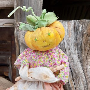 May include: A handmade pumpkin doll with a smiling face, green embroidered eyes, and a green stem. The doll wears a pink floral print dress and holds a white cloth. The background is a weathered wooden surface.