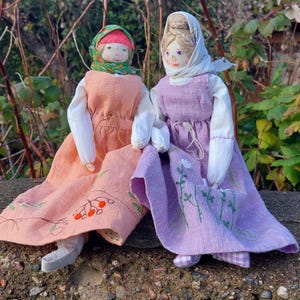 Peut inclure: Deux poupées en tissu faites à la main portant des robes et des couvre-chefs. La poupée de gauche porte une robe pêche avec des fleurs brodées et des baies rouges. La poupée de droite porte une robe lavande avec des fleurs brodées et une bordure en vichy blanche.