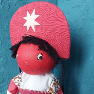 May include: A red fabric doll with black hair, a white star on a red hat, and a white dress with floral accents.