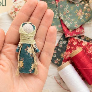 Può includere: Una bambola in miniatura in tessuto con un vestito color verde acqua a motivi floreali gialli e bianchi, un copricapo verde chiaro e una testa in tessuto beige semplice. La bambola è decorata con sottili spaghi marrone chiaro legati in vita.