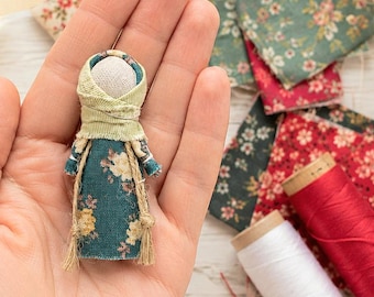 Puede incluir: Una muñeca de tela en miniatura con un vestido color verde azulado con estampado floral amarillo y blanco, un pañuelo de cabeza verde claro y una cabeza de tela beige lisa. La muñeca está adornada con finos cordeles marrones atados en la cintura.