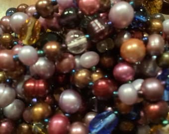 May include: A close-up of a strand of pearls and beads in various colors, including pink, purple, brown, and white. The beads are arranged in a random pattern, creating a visually appealing and textured look.