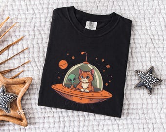 May include: Black t-shirt with a graphic of a cat and an alien in a flying saucer. The cat is orange and white, and the alien is green. The flying saucer is orange and has a clear dome. The background is a dark blue with stars.