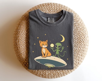 May include: A dark gray t-shirt with a graphic print of a cat and an alien on a flying saucer. The cat is orange and the alien is green. The background is a dark blue with stars.