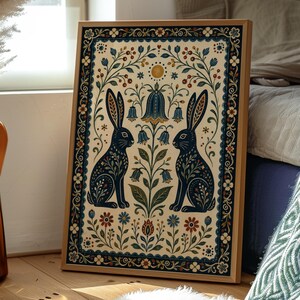 Può includere: Una stampa rettangolare incorniciata raffigurante due conigli blu scuro uno di fronte all'altro, circondati da motivi floreali simmetrici, fiori blu a forma di campana e un piccolo sole giallo su sfondo color crema con un bordo decorativo.