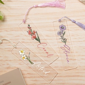 May include: Three clear acrylic bookmarks with floral designs and names "Astrid", "Coral", and "Gloria" written on them. Each bookmark has a tassel attached.