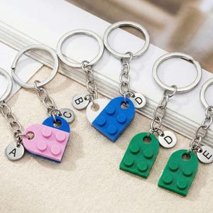 Può includere: Portachiavi con anelli e catene argentate, con ciondoli colorati a forma di mattoncino. I ciondoli sono rosa, blu e verdi, con ciondoli con lettere argentate. I portachiavi sono disposti su una superficie bianca.