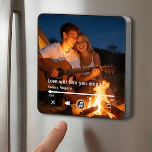May include: Square magnet with a photo of a couple by a campfire, with a guitar. The text "Love will turn you around" by Kenny Rogers is displayed. The magnet is attached to a stainless steel refrigerator.