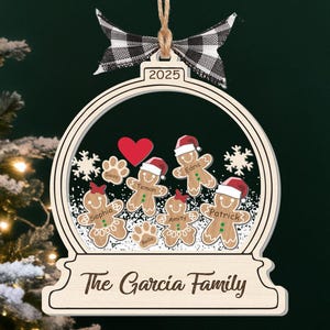 May include: Wooden Christmas ornament shaped like a snow globe, featuring gingerbread people with names, a red heart, and paw prints. The ornament has the text "The Garcia Family" and the year "2025". A black and white checkered bow is at the top.