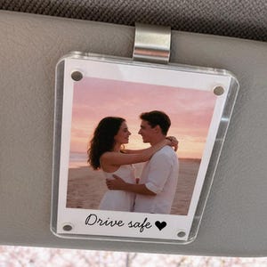 May include: A clear acrylic car visor photo holder with a photo of a couple embracing on a beach. The text "Drive safe" and a heart are printed below the photo. The visor is attached to a car's interior.