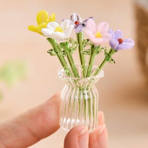 May include: A small glass vase with a clear, textured design holds a bouquet of colorful glass flowers. The flowers are yellow, white, pink, purple, and blue.