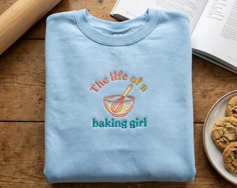 May include: Light blue sweatshirt with the embroidered text "The life of a baking girl" above a whisk and bowl design. The sweatshirt is folded on a wooden surface, with a rolling pin and a cookbook in the background.