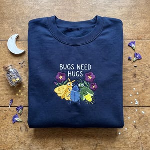 May include: A navy blue crewneck sweatshirt featuring an embroidered design of three colorful insects hugging. The text "BUGS NEED HUGS" is stitched in white above the insects, which are surrounded by small purple flowers on a dark fabric background.