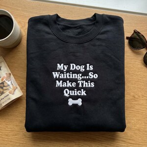 May include: Black sweatshirt with white embroidered text that reads "My Dog Is Waiting...So Make This Quick" and a bone graphic. The sweatshirt is folded on a wooden surface, with a cup of coffee and dog treats visible.